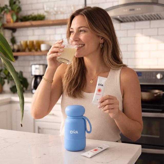 A woman drinking in her kitchen, the OYA bottle beside her